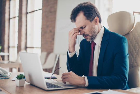 Exhausted boss is holding his nose bridge with closed eyes, he is tired and depressed. He is broker, he is wearing suit with tie and is sitting at his work place