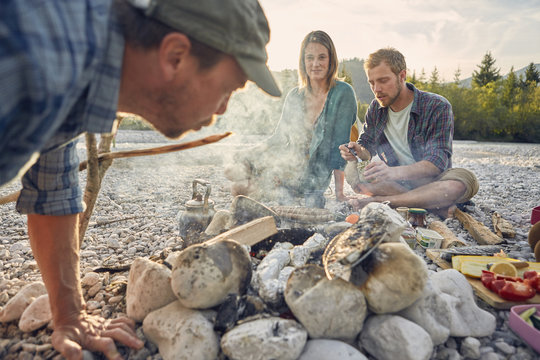 Adults sitting around campfire preparing food and blowing on embers