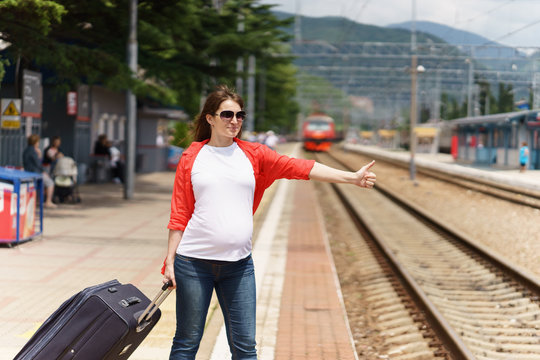 young european pregnant woman with heavy baggage trying to stop train on railway station for traveling at sunny day