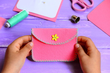 Obraz premium Child made a felt purse. Small child holds a pink felt purse in his hands. Craft supplies on a wooden table. Fun and easy sewing project for kids