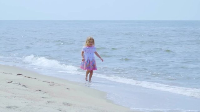 Adorable little girl  in a dress runs  jumping on the beach.  Child enjoys playing on the seashore running on the water with pleasure. splashing in sea waves.