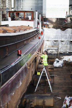 Worker on step ladder checking boat in shipyard workshop