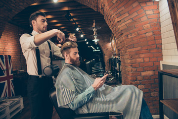 Side view of handsome young red bearded man browsing at his pda, while getting a haircut by classy dressed barber shop stylist