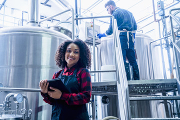 Mid adult woman in brewery wearing apron holding digital tablet looking at camera smiling