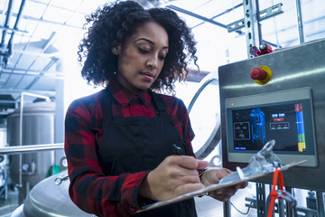 Mid adult woman in brewery monitoring computer equipment, looking down writing on clipboard