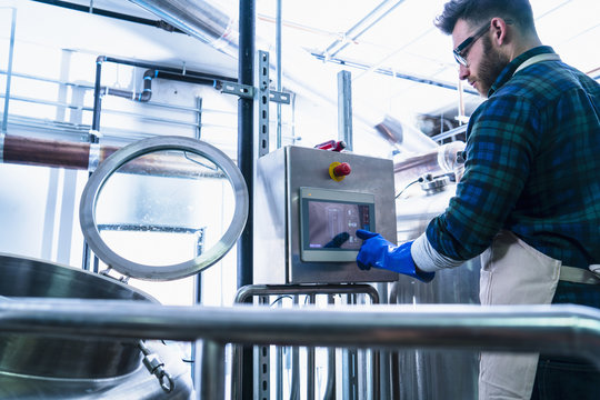Young man in brewery wearing apron and protective gloves using touch screen computer equipment