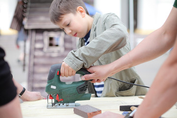 DIY teacher or father teaching son tinkering, using jig saw. Classes for young carpenter polish wood board form using jigsaw. Toy making. Dreaming of future profession. child woodcarving lesson