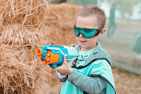 Small Preschooler Boy With Blaster Prepare For Attack And Play With Friends In Protective Glasses. Excited Child With Darts Toy Gun On The Play Field