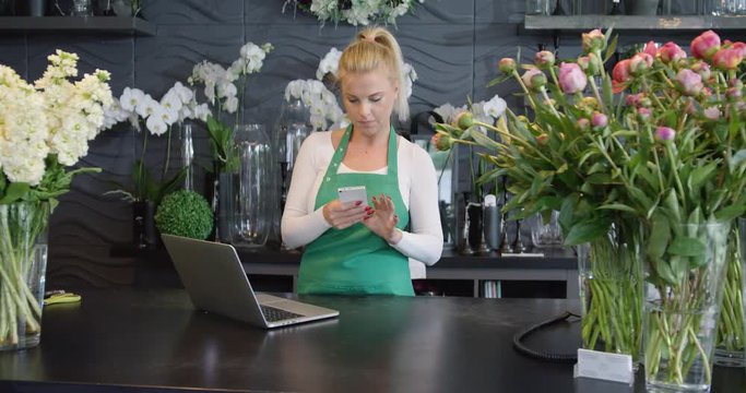 Young female florist wearing uniform and working in floral shop while using smartphone and standing behind counter.
