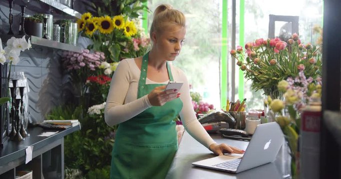 Side view of young woman in green apron standing with smartphone in hands behind counter and browsing laptop.
