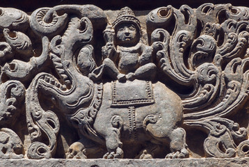 Fototapeta na wymiar Ancient Hindu god sitting on myth lion creature, sculpured stone relief carvings from the 12th century temple's wall, Halebidu town of Karnataka. Old Indian artwork
