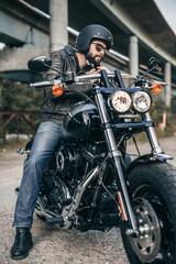 Outdoor lifestyle portrait of handsome biker man sitting on a motorcycle. Biker man wearing jeans and leather jacket sitting on motorbike.