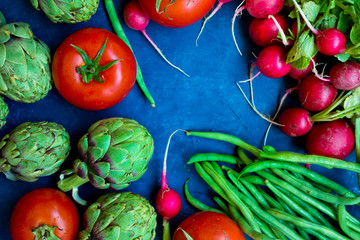 Variety of fresh colorful organic vegetables green beans, tomatoes, red radish, artichokes on dark blue background, copy space, conceptual, healthy food, vegetarian, vegan