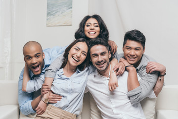 young cheerful multiethnic friends embracing while sitting on sofa at home