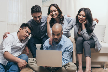 Fototapeta premium young smiling multiethnic friends looking at laptop while sitting on sofa at home