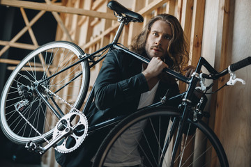 Plakat Stylish bearded young man with long hair carrying bicycle on shoulder