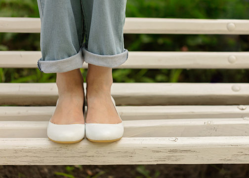 Female legs in blue jeans and white shoes on a white bench.