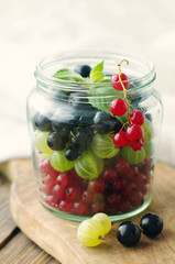 Fresh berries of a gooseberry, black and red currant n a glass jar on a wooden board. Summer harvest of berries
