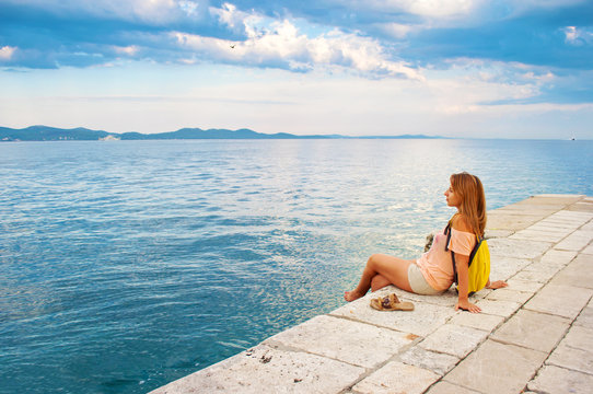 One young girl with a backpack sitting peacefully sideways near the sea shore looking wistfully at the hill range in the distance, hands leaning against stone pavement, legs crossed. Zadar, Croatia