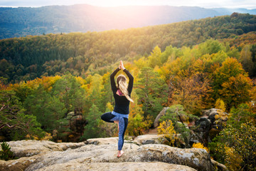 Attractive young female is practicing yoga and doing asana Vrikshasana on the top of the high rocky mountain in the evening. Autumn forests, rocks and hills on the background