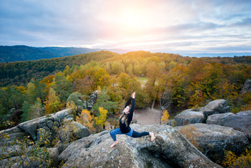 Flexible fit woman is practicing yoga and doing asana Virabhadrasana 1 on the top of the huge boulder in the evening. Autumn forests, rocks and hills on the background