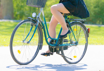 Fototapeta premium Girl on bicycle wearing on black short dress. Young Woman riding along road on green spring outdoor Park. Sporty young girl riding a bicycle on a sunny morning