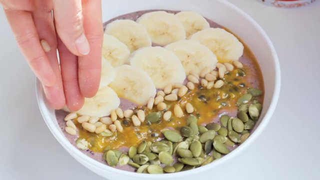 Close up of female hands decorating big blueberry smoothie bowl with banana, passion fruit and cedar nuts. Concept of healthy food and lifestyle