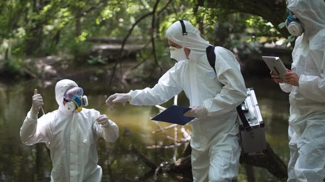 Biochemist in suit taking sample of water in river in test tube while investigating nature with coworkers.