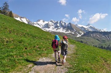 Fototapeta na wymiar Zwei Kinder wandern Hände haltend auf Wanderweg in den Schweizer Bergen auf Wanderweg. Grüne Wiese und Schneeberge im Hintergrund
