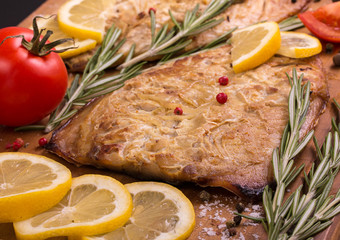 Baked mackerel with slices of lemon, rosemary, tomatoes, garlic, salt, red and black pepper on wooden cutting board, close up.