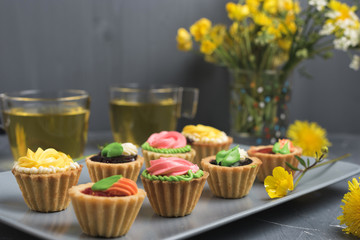 Colorful cupcakes on grey plate and table. Cakes with cream and jam. Cup of green tea and yellow flowers on a background