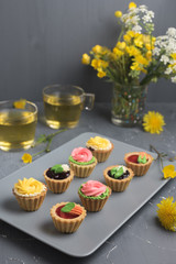 Colorful cupcakes on grey plate and table. Cakes with cream and jam. Cup of green tea and yellow flowers on a background