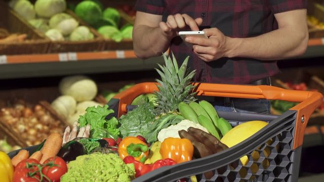 Young man checking shopping list on his smartphone at the hypermarket. Close up of male hands typing on touchscreen of the cellphone. Guy pointing on different fruits and vegetables in his grocery