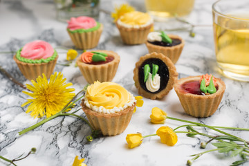 Colorful cupcakes on grey marble table. Cakes with cream and jam. Cup of green tea and yellow flowers on a background