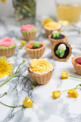 Colorful cupcakes on grey marble table. Cakes with cream and jam. Cup of green tea and yellow flowers on a background