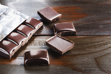 Bitter, dark chocolate bar in foil and broken cubes on wooden background
