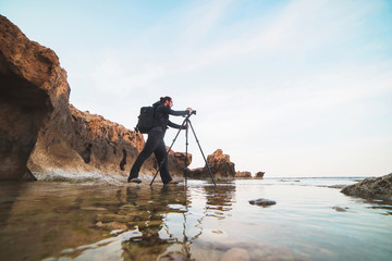 Young stylish photographer making photos of sea with the camera on a tripod - obrazy, fototapety, plakaty