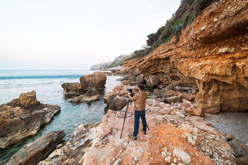 Young male photographer standing on rocks with tripod and making photos of sea