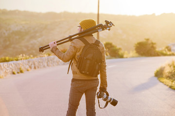 Fototapeta premium Young photographer walking on desert road with tripod on his shoulder and camera in hand