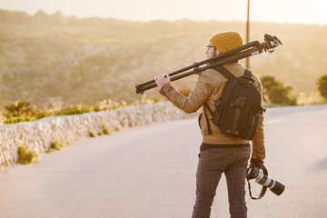 Young photographer walking on desert road with tripod on his shoulder and camera in hand
