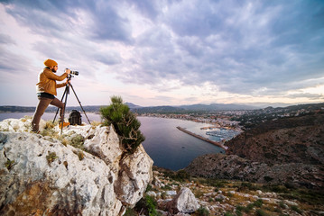 Young attractive photographer making photos with camera on tripod at sunset