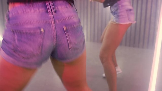 Two grils dancing club. Girls in denim shorts on lamp light background. Close up of sexy girls shaking buttocks in slow motion. Two women dancing at disco party. Women shaking hips in disco club
