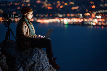 Young attractive photographer sitting on rock and working on laptop with view of sea and mountains