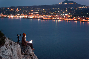 Young attractive photographer sitting on rock and working on laptop with view of sea and mountains