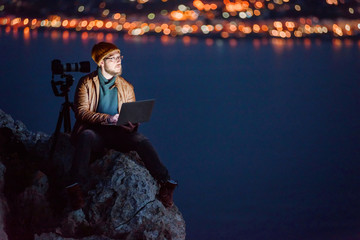 Young dreamy photographer sitting on precipice and working on laptop with view of sea and mountains