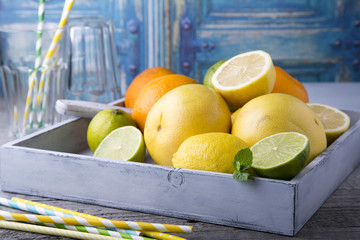 Assorted citrus fruits with colorful cocktail straws in a light wooden box on a gray wooden table opposite the blue old door