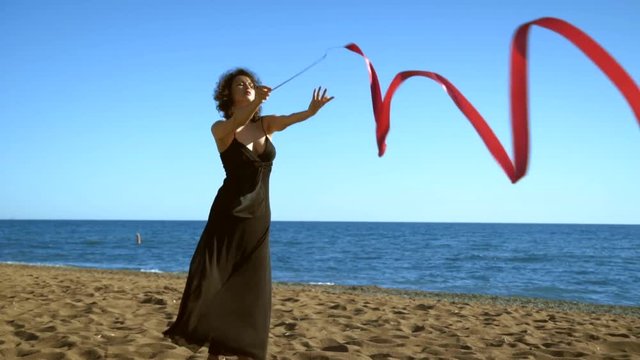 A young woman in a black dress is standing on the sand and doing a drawing of a red ginastic ribbon