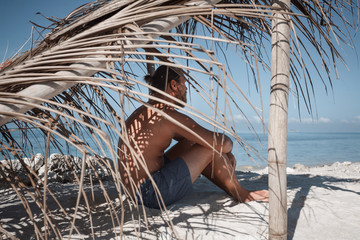 Young man traveler sitting on beach in the shade of hovel made of palm leaves