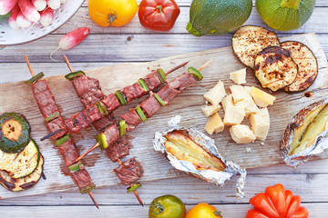 Top view of bbq beef or pork on skewers, baked potato, eggplant, cheese on wooden board served with fresh vegetables. 