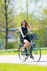 Portrait view of girl on bicycle wearing on black short dress. Young happy Woman riding along road on green spring  outdoor Park. Sporty young girl riding a bicycle on a sunny morning, view from face